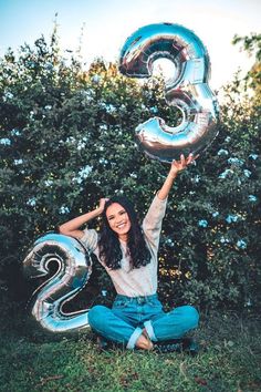 a woman sitting in the grass with her hands up and number three balloons above her head