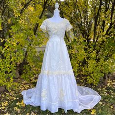 a white wedding dress on display in front of some trees and bushes with yellow leaves