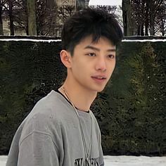 a young man standing in front of a green wall with snow on the ground and trees behind him