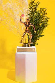 a gold statue holding a frisbee on top of a white pedestal next to some plants