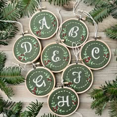 six embroidered christmas ornaments hanging on a wooden table with evergreen leaves and pine cones around them