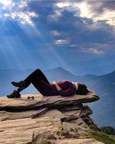 a woman laying on top of a large rock next to a mountain under a cloudy sky