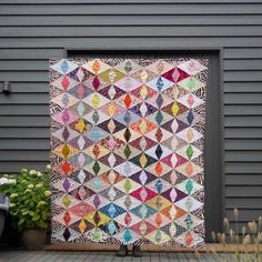 a large quilt hanging on the side of a building with potted plants next to it