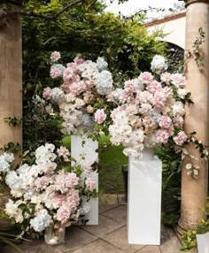 three white vases with pink and white flowers in them on a stone walkway next to trees