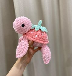 a small pink crocheted stuffed animal with a green leaf on its head is held up in front of a white curtain