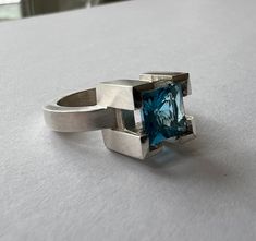 A princess-cut Blue Topaz Ring set in a well protected, cast v-shape claw ring.  The bold, geometric high setting is a design feature of this ring.  This sterling silver ring has a brushed finish with a high polished top and contrasting burnished edges. AVAILABLE - IN STOCK  Size - 5 - this ring could be sized up to a 5.25. Other sizes and different gemstones (peridot, amethyst, citrine and garnet) may be available on a made to order basis.  Processing time would be 4-6 weeks.  Please inquire. Architectural Aesthetic, Claw Ring, Geometric Ring, Blue Band, Blue Topaz Ring, Topaz Ring, Princess Cut, Gemstone Ring, V Shape