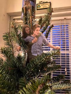 two people standing in a christmas tree with their arms around each other and pointing at something