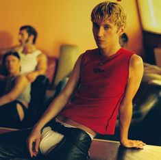 three young men sitting in a living room with one wearing a red shirt and black pants