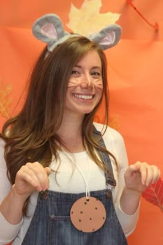 a woman wearing bunny ears and overalls holds out her hand to the camera while standing in front of an orange backdrop