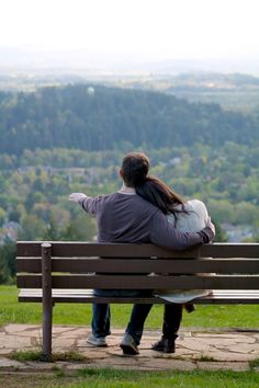 a man and woman sitting on a bench looking out at the valley in the distance