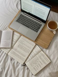 an open laptop computer sitting on top of a bed next to books and a cup of coffee