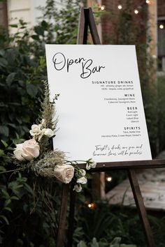 an open bar sign with flowers and greenery on it in front of a building