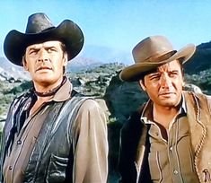 two men wearing cowboy hats standing next to each other in front of a mountain range