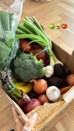 a box filled with lots of vegetables on top of a wooden floor next to a person's hand