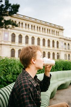 a young man sitting on a bench drinking from a white cup in front of a building