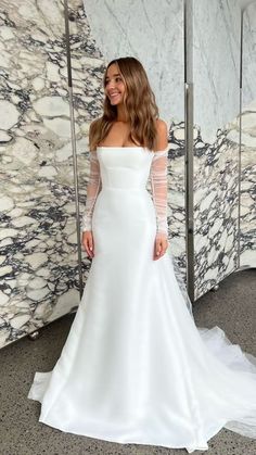 a woman standing in front of a marble wall wearing a white dress with sheer sleeves