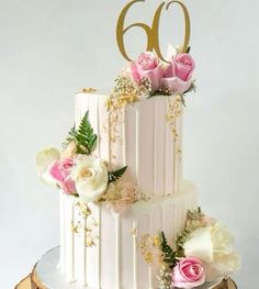 a white wedding cake with pink flowers and gold numbers on the top is sitting on a table