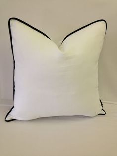 a white pillow with black piping on the front and back side, sitting against a wall