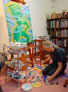 an older woman is painting on the floor in front of a bookshelf full of art supplies