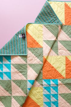 an orange and blue quilt is laying on top of a pink background with a white tag in the middle