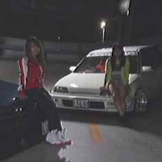 two women sitting on suitcases in front of a white car and another woman standing next to her