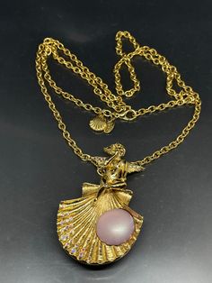 This vintage Kirks Folly necklace is a stunning piece of jewelry that will complement any outfit. The necklace features a gold-tone chain with a lobster closure and a pendant in the shape of a shell that also can be used as a brooch. The shell is adorned with Aurora Borealis crystals and a purple imitation pearl. The pendant also has a beautiful cherub or angel figure with crystal accents. This necklace is 30 inches long, making it the perfect length to wear with any neckline. It is a pre-owned vintage piece of jewelry that has been well-maintained and signed by the brand. Add this unique and glamorous necklace to your collection today! Elegant Gold Necklace With Brooch, Evening Brooch Pendant Necklaces, Pendant Necklaces With Brooch For Evening, Vintage Necklace With Large Pendant For Evening, Vintage Evening Necklace With Large Pendant, Vintage Large Pendant Necklace For Evening, Gold Pendant Necklace With Brooch, Vintage Gold-tone Necklace With Large Pendant, Angel Cherub