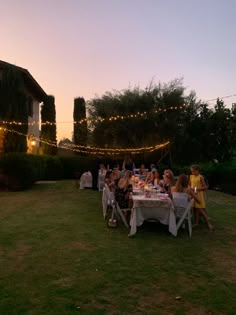 a group of people sitting around a table outside at night with lights strung across the lawn