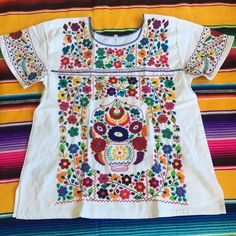 an embroidered mexican shirt with flowers and leaves on the chest, sitting on a colorful striped blanket