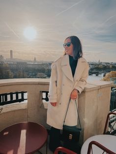 Cream coat and jeans standing against a wall in Paris with sun in background.