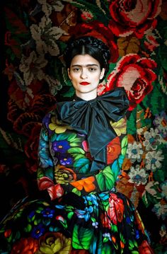 a woman sitting in front of a floral wallpaper wearing a black bow neck dress