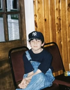 a young boy in overalls and a new york yankees cap sitting on a chair