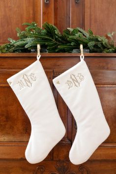 two white christmas stockings hanging from a wooden mantle with evergreen branches and pine cones on top