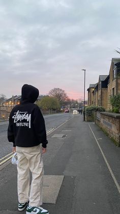 a person standing on the sidewalk with their back to the camera, wearing a black hoodie and white pants