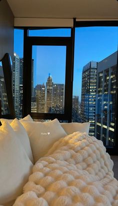 a bed with white pillows in front of large windows and cityscape at night