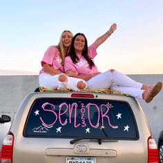 two women in pink shirts sitting on the back of a silver car with their arms up