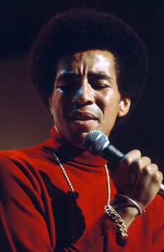 a man holding a microphone in his right hand while wearing a red shirt and chain around his neck