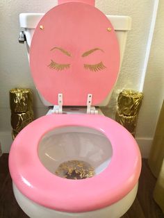 a pink and white toilet with eyelashes painted on the lid, next to gold decorations