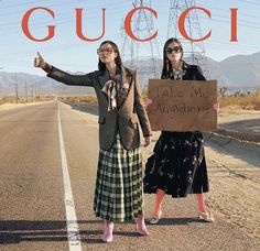 two women standing in the middle of an empty road holding a sign that says gucci