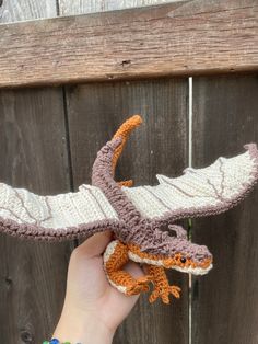 a hand is holding a crocheted dragon ornament in front of a wooden fence