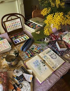 an assortment of art supplies on a table with yellow flowers in the vase and other items