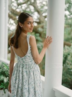 a woman in a dress standing on a porch next to a white column and looking off into the distance