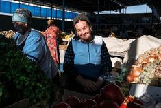 two men in aprons are at an open air market with vegetables and fruit for sale