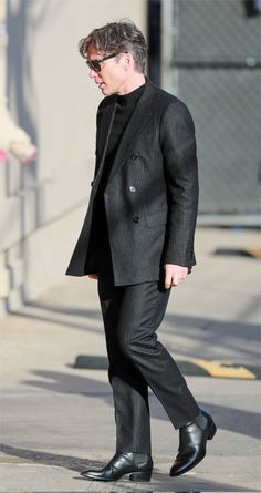 a man in a black suit and sunglasses walking down the street with his hand in his pocket