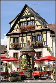 an old building with flowers in the window boxes on it's roof and some tables under umbrellas