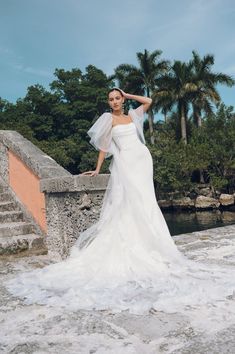 a woman in a white wedding dress standing on a stone bridge with palm trees behind her