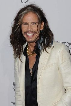 a man with long hair wearing a white suit and pearls on his necklace smiles at the camera