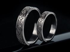 two wedding rings are shown on a black surface, one is engraved with an intricate design