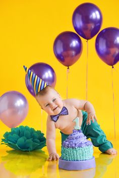 a baby wearing a birthday hat and sitting on top of a cake with balloons in the background