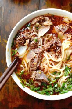 a bowl filled with noodles, meat and vegetables on top of a wooden table next to chopsticks