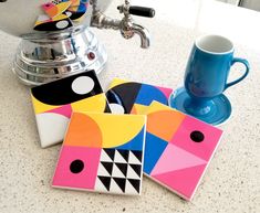 some colorful coasters sitting on top of a counter next to a blue cup and silver faucet